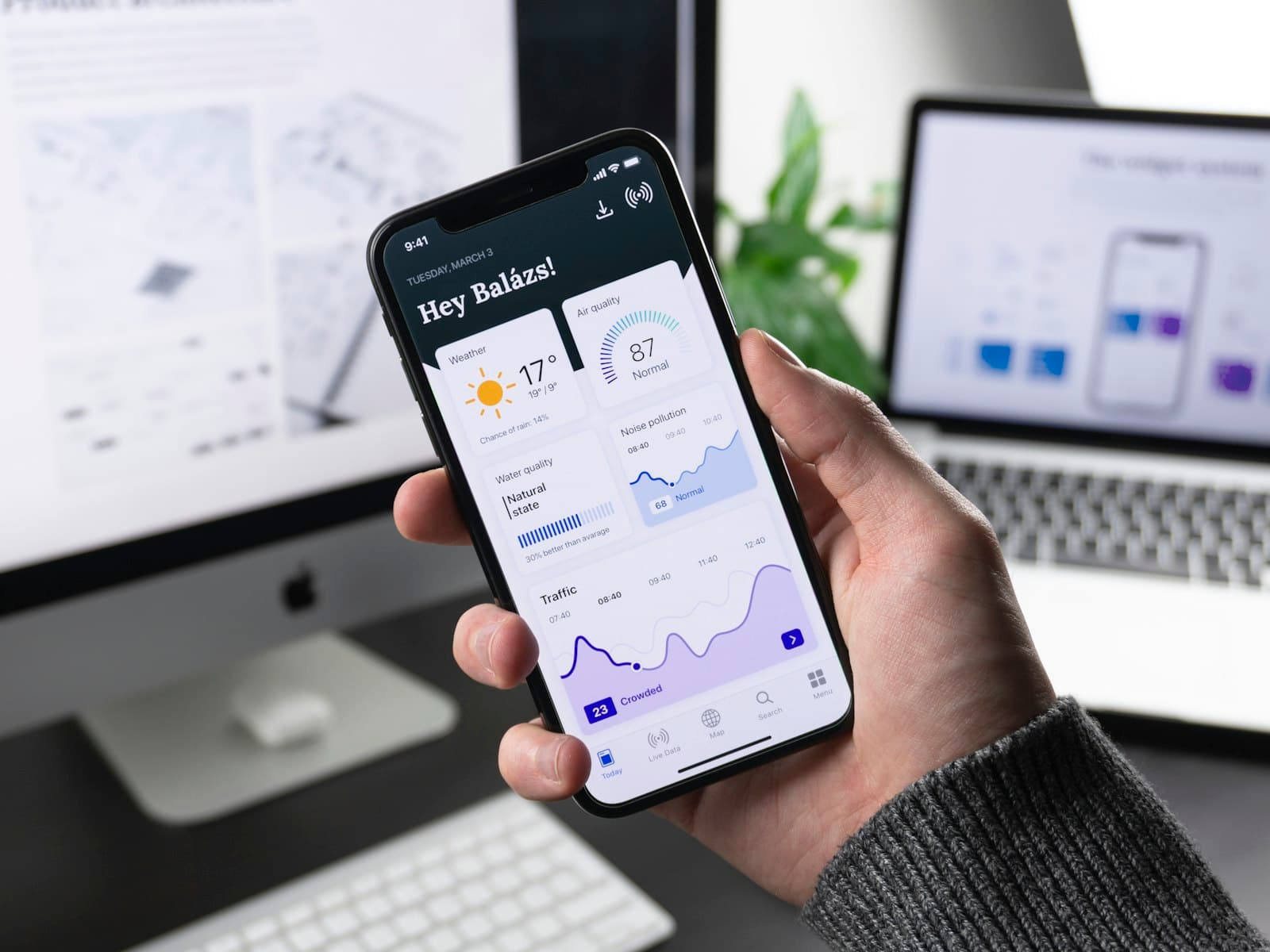 Hand hält ein Smartphone mit App-Dashboard vor einem Desktop-Bildschirm
